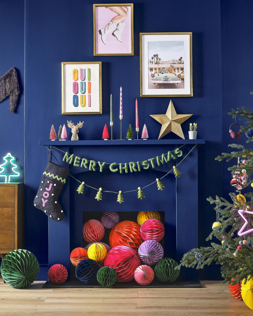 Decorative Christmas scene with colorful ornaments, a 'Merry Christmas' banner, and framed pictures on a blue wall.