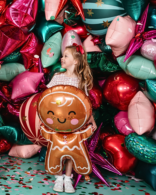 Child surrounded by colorful balloons, including a gingerbread man balloon, on a green surface with confetti.