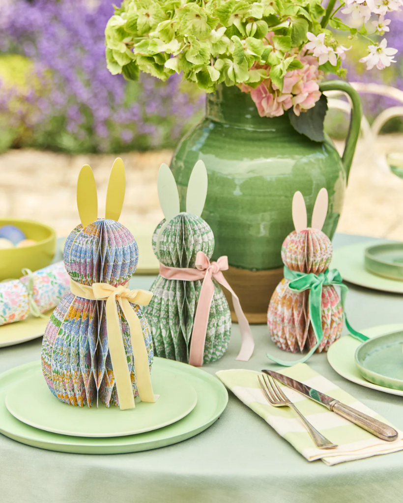 Decorative paper bunnies on a table with a green vase and flowers in the background.