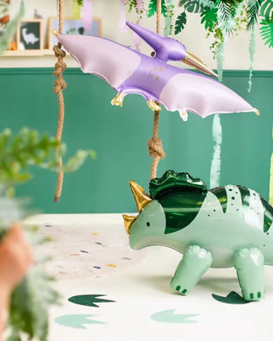 A purple pterosaur balloon with rope handles hangs above a green and white dinosaur figurine on a white surface, set against a teal wall with hanging plants.