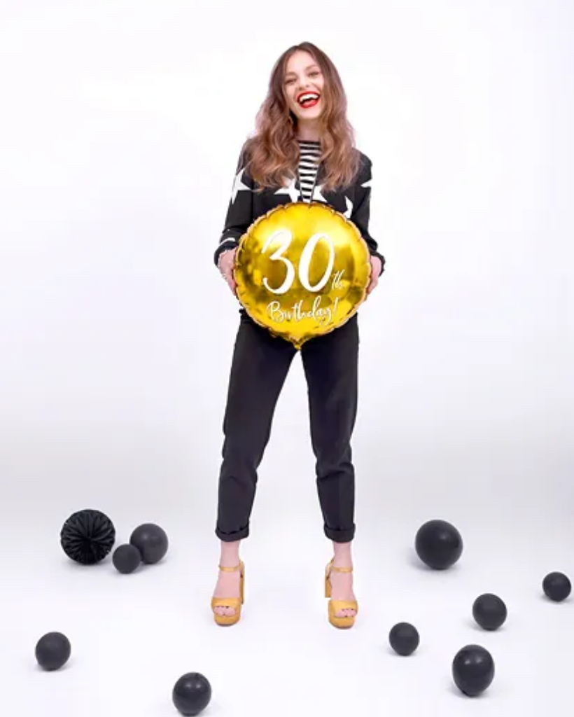 Woman holding a gold '30th Birthday' balloon with black and gold balloons on the floor.