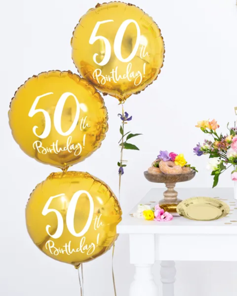 Three yellow '50th Birthday!' balloons on a white background with a table and flowers in the background.