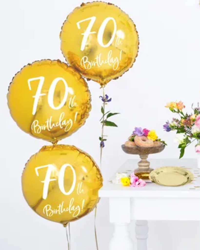Three gold '70th Birthday!' balloons with a cake and flowers on a table in the background.