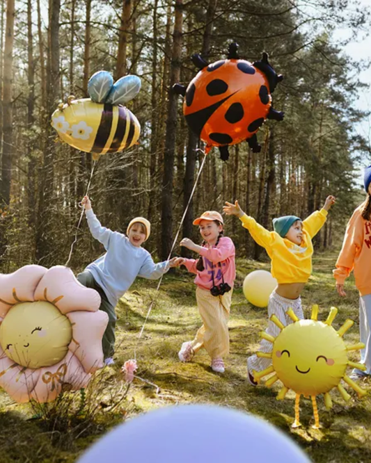 Children playing with colorful balloons shaped like a bee, ladybug, sunflower, and sun in a forest.