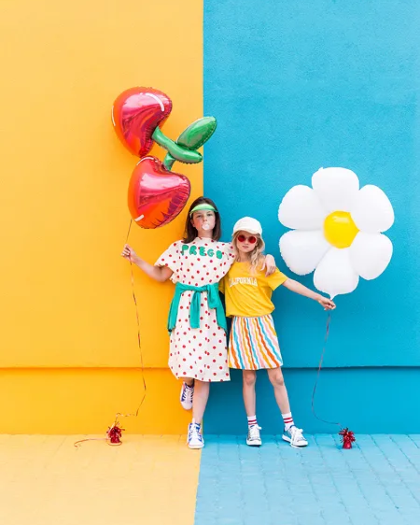 Two children holding colorful balloons against a yellow and blue wall.