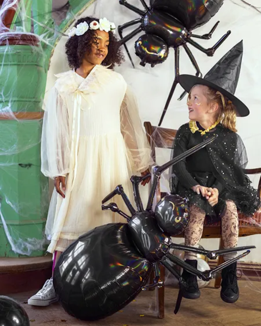 Two children in Halloween costumes standing next to a large black spider balloon.