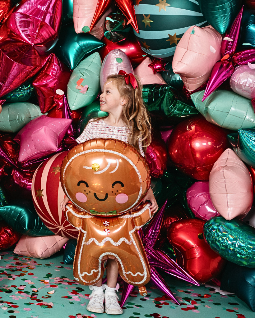 Child surrounded by colorful balloons, including a gingerbread man balloon, on a green surface with confetti.