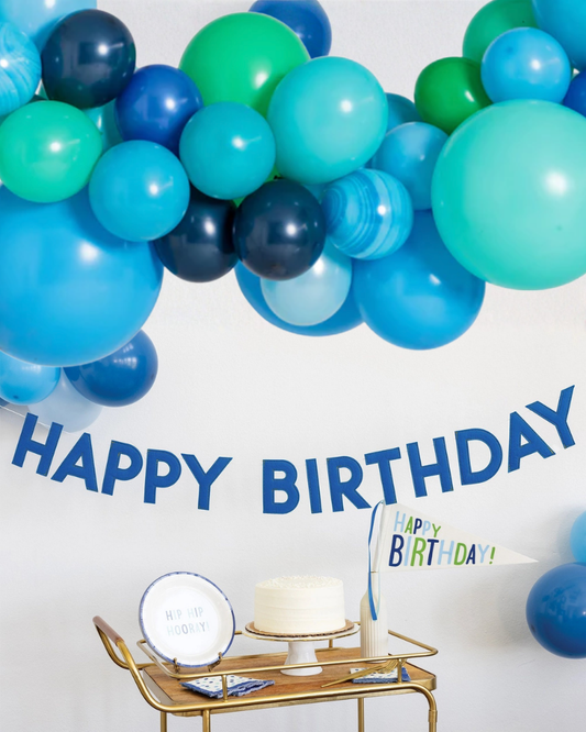 Blue and green balloon arch with 'Happy Birthday' banner and cake on a table.
