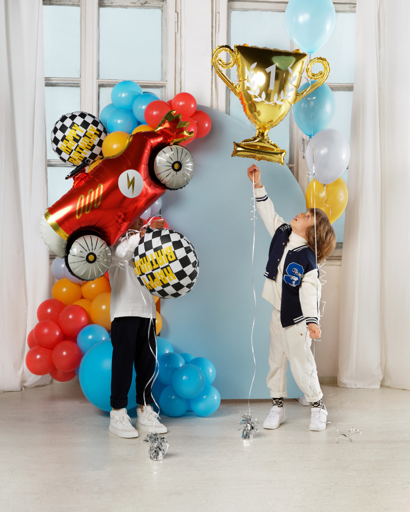 Two children playing with colorful balloons and a trophy in a room with large windows.