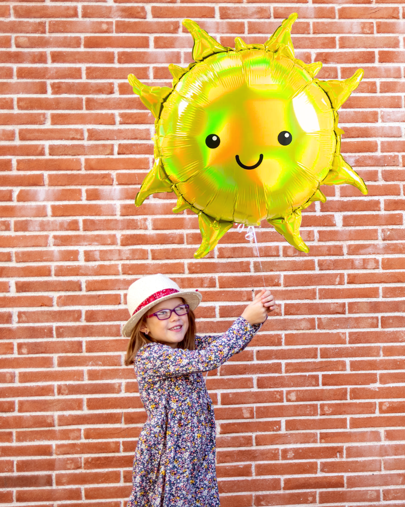 Child holding a sun-shaped balloon against a brick wall