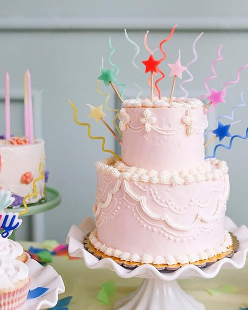 Two-tiered pink cake with colorful candles and decorations on a white stand against a light blue background.