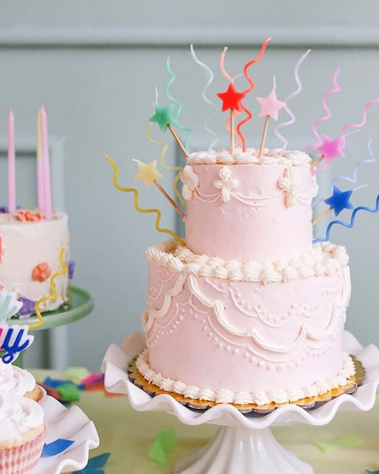 Two-tiered pink cake with colorful candles and decorations on a white stand against a light blue background.