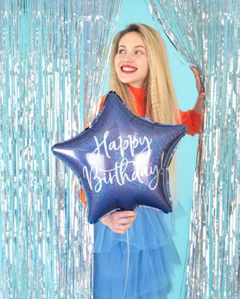 Woman holding a blue 'Happy Birthday' balloon in front of a silver tinsel curtain.