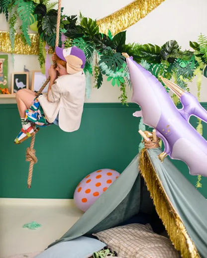A child wearing a purple and beige cap, cream jacket, and colorful patterned shorts hangs from a rope swing in a decorated room with a green wall, gold garland, a large purple dinosaur balloon, and a small play tent.