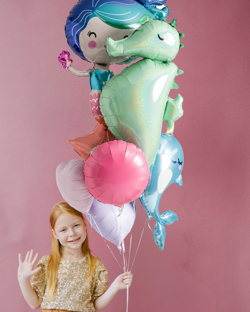 A young girl with long red hair smiles while holding a cluster of mermaid-themed foil balloons against a solid pink background. The balloons include a seahorse, a mermaid, a blue dolphin, and pink and lavender accents.