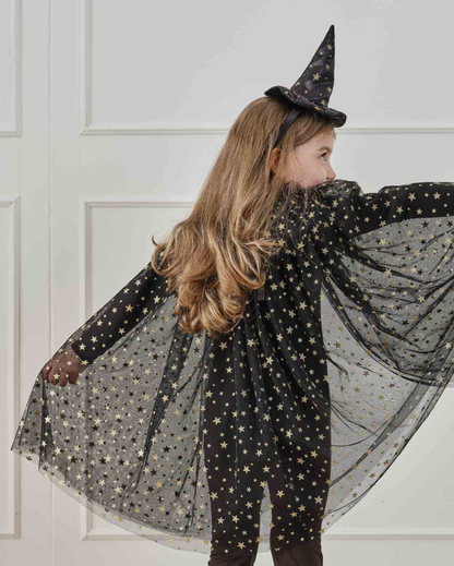 A child with long blonde hair is shown from the back wearing a black witch costume cape and matching pointed hat, both decorated with gold star patterns. They are posing with her arms outstretched against a white paneled wall.