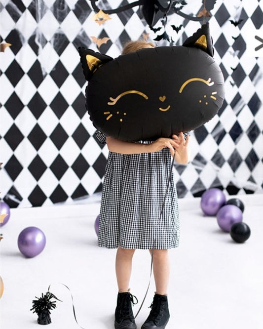 Child holding a black cat-shaped foil balloon against a checkered background with Halloween decorations.