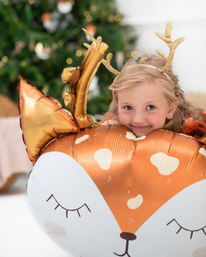 Child wearing reindeer antlers with a large inflatable deer foil balloon in front of a decorated Christmas tree.
