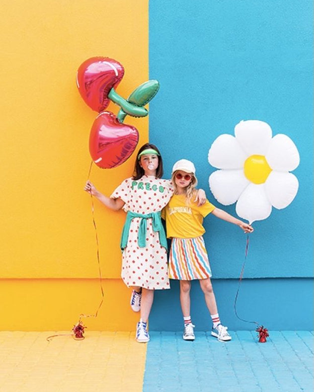 Two children holding balloons against a colorful wall with a yellow heart and red flower balloon and a white daisy balloon.