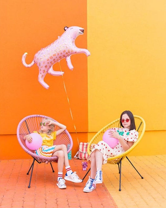 Two people sitting on colourful chairs with inflatable balloons and a pink leopard foil balloon against a two-tone orange and yellow wall.