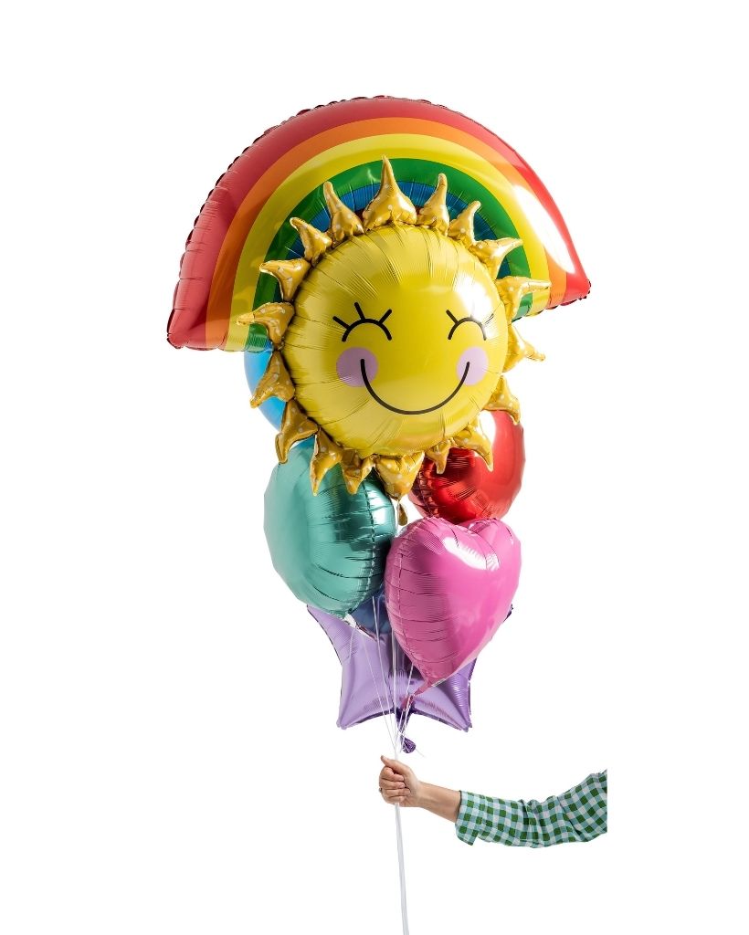 A bouquet of foil balloons in the shapes of a sun and a rainbow with smiling faces, held by a hand. The balloons are multicolored with shades of yellow, red, green, and purple.