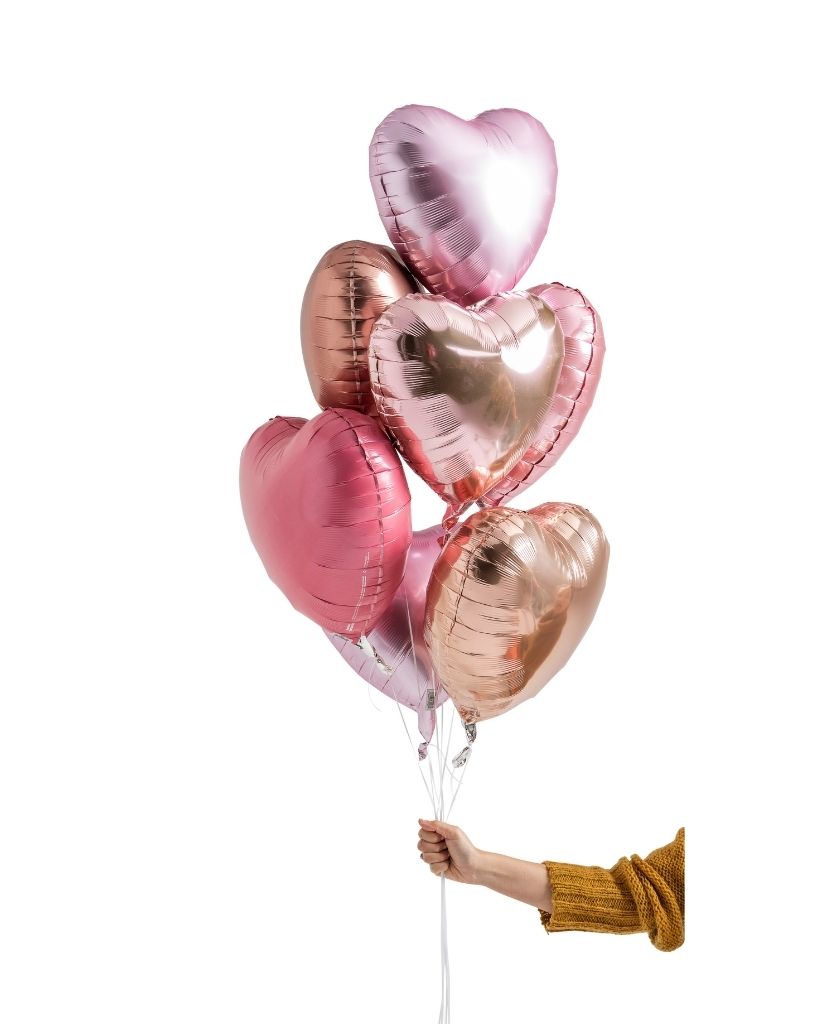 A set of heart-shaped foil balloons in shades of pink and rose gold, held by a person against a white background.